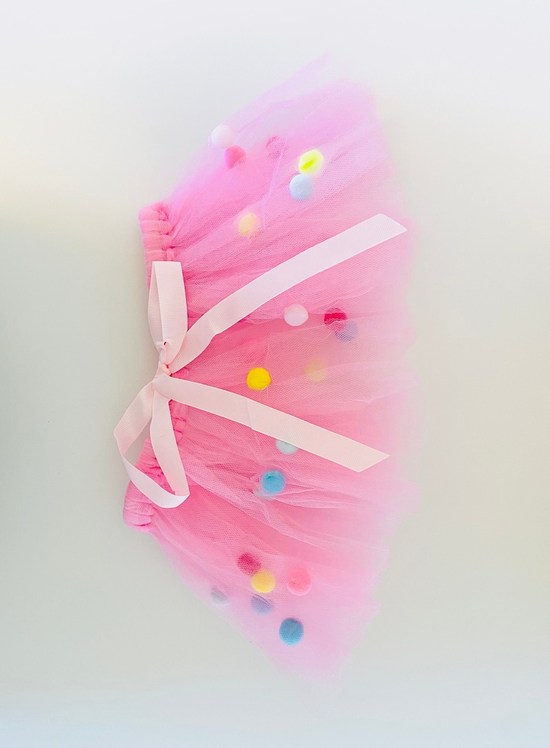 May include: A pink tulle tutu skirt with a white ribbon and colorful pom-poms.
