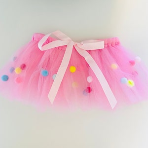 May include: A pink tulle tutu skirt with a white satin bow and colorful pom-poms.