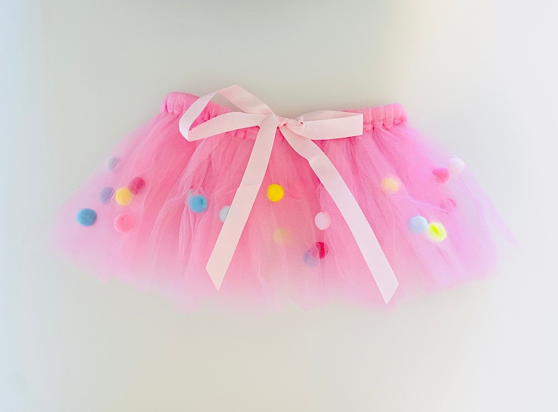May include: A pink tulle tutu skirt with a white satin bow and colorful pom-poms.