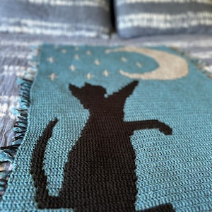 May include: A crocheted blanket featuring a blue background with a black cat silhouette. The cat is gazing upwards at a crescent moon with white stars.
