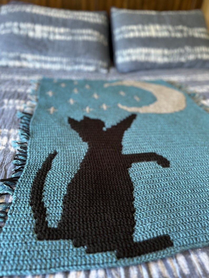 May include: A crocheted blanket featuring a blue background with a black cat silhouette. The cat is gazing upwards at a crescent moon with white stars.