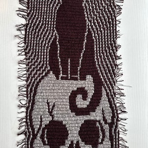 May include: A crocheted wall hanging with a skull and a cat design. The skull is gray and the cat is maroon. The background is a wavy pattern in gray and maroon.
