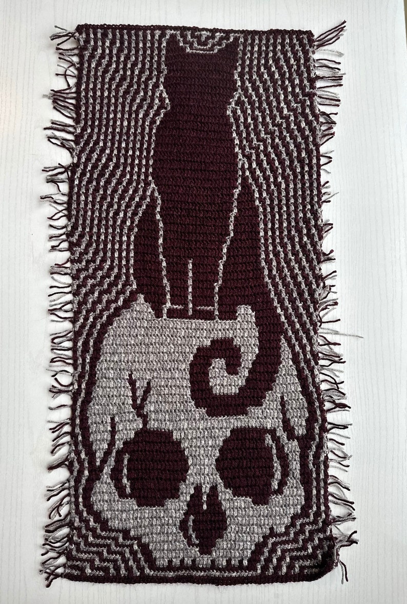 May include: A crocheted wall hanging with a skull and a cat design. The skull is gray and the cat is maroon. The background is a wavy pattern in gray and maroon.