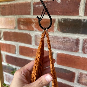 May include: A brown crocheted plant hanger with two loops hanging from a black S-shaped hook.