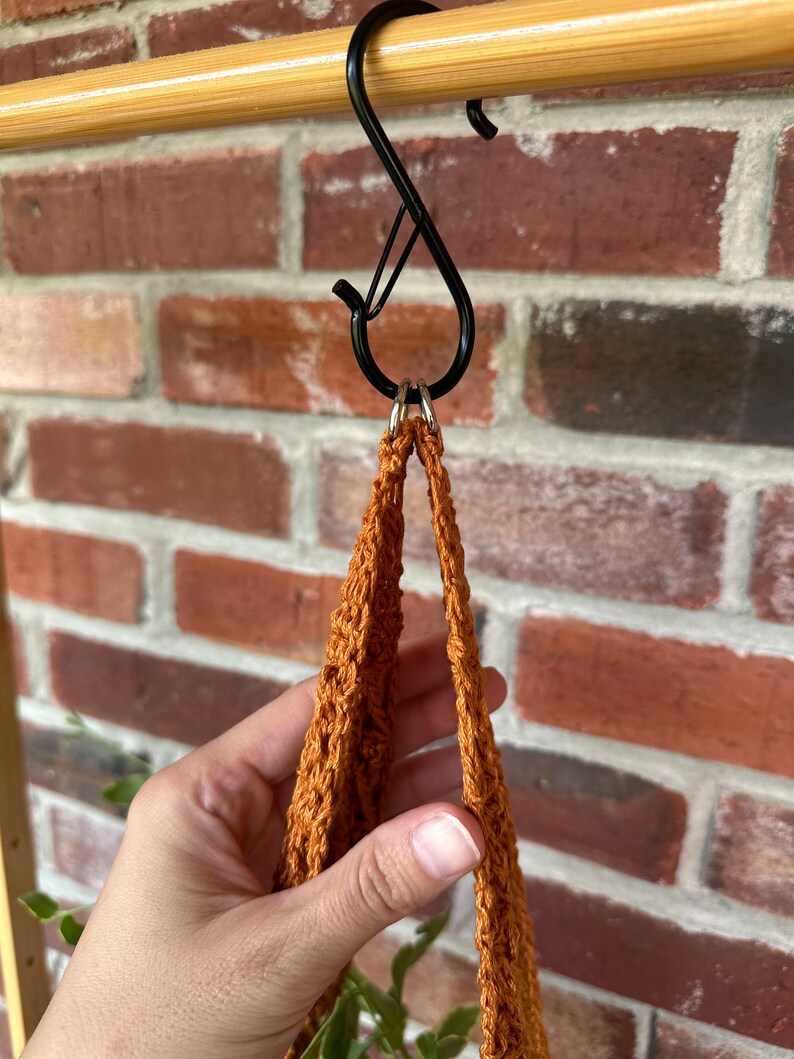 May include: A brown crocheted plant hanger with two loops hanging from a black S-shaped hook.