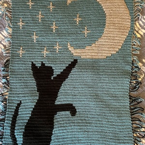 May include: A crocheted blanket featuring a black cat silhouette reaching up to a crescent moon with stars on a teal background.