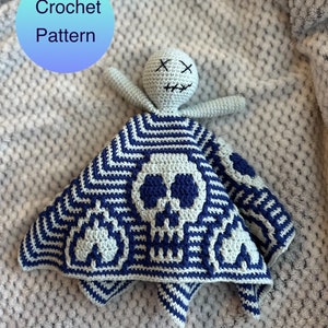 Puede incluir: Una manta de bebé tejida a crochet con un diseño de calavera en azul y gris. La manta está doblada y una muñeca de crochet con una cara bordada está encima. El texto "Crochet Pattern" está escrito en un círculo azul y verde.