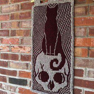 May include: A crocheted wall hanging featuring a black cat standing on top of a skull. The cat and skull are both made of yarn and have a textured appearance. The background of the hanging is a light gray color.
