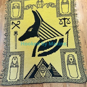 May include: A yellow and black throw blanket featuring an Anubis design. The blanket has a repeating geometric border and fringe. The Anubis image is in the center, with other Egyptian symbols around it. The text "Hooked by Maxine" is in the image.