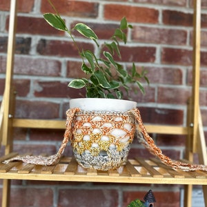 May include: A small potted plant with green leaves in a white pot, sitting on a wooden shelf. The pot is wrapped in a crocheted cover with orange, yellow, and gray stripes.