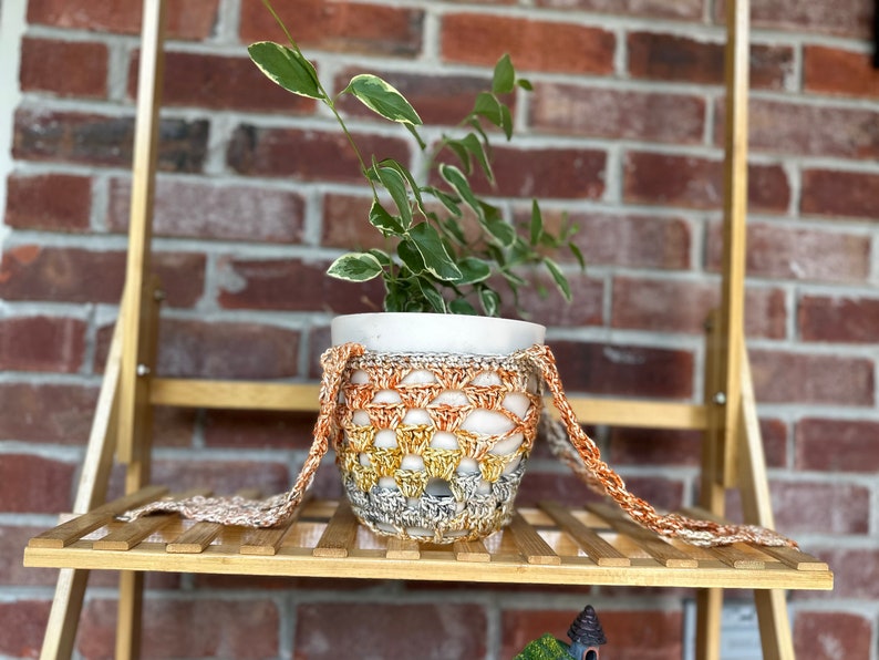 May include: A small potted plant with green leaves in a white pot, sitting on a wooden shelf. The pot is wrapped in a crocheted cover with orange, yellow, and gray stripes.
