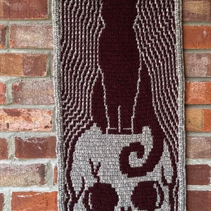 May include: A crocheted wall hanging featuring a black cat standing on a skull. The cat and skull are made with maroon yarn, while the background is a gray yarn.