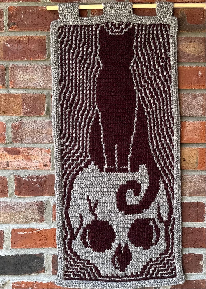 May include: A crocheted wall hanging featuring a black cat standing on a skull. The cat and skull are made with maroon yarn, while the background is a gray yarn.