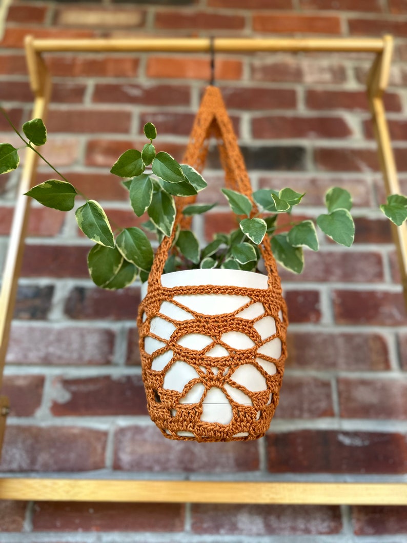 May include: A terracotta crocheted plant hanger with a white ceramic pot and a green plant inside. The hanger is hanging from a wooden shelf.