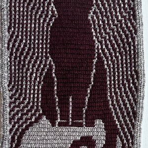 May include: A close-up of a crocheted tapestry featuring a stylized image of a person standing on a platform. The tapestry is made with brown and gray yarn, creating a textured effect.