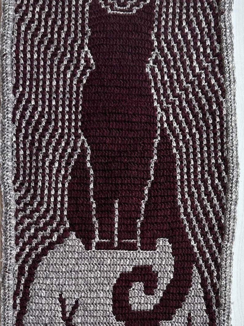 May include: A close-up of a crocheted tapestry featuring a stylized image of a person standing on a platform. The tapestry is made with brown and gray yarn, creating a textured effect.