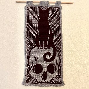May include: A crocheted wall hanging featuring a black cat silhouette standing on top of a gray skull with a swirl design. The background is a gray and white striped pattern.