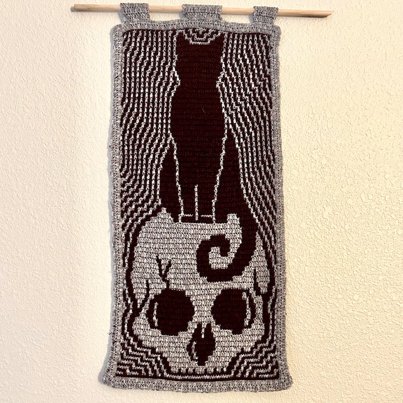 May include: A crocheted wall hanging featuring a black cat silhouette standing on top of a gray skull with a swirl design. The background is a gray and white striped pattern.