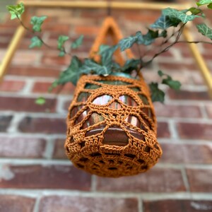 May include: A brown crocheted hanging planter with a green plant inside. The planter is hanging from a wooden frame against a brick wall.
