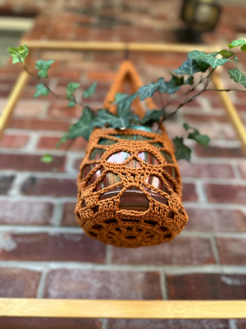 May include: A brown crocheted hanging planter with a green plant inside. The planter is hanging from a wooden frame against a brick wall.