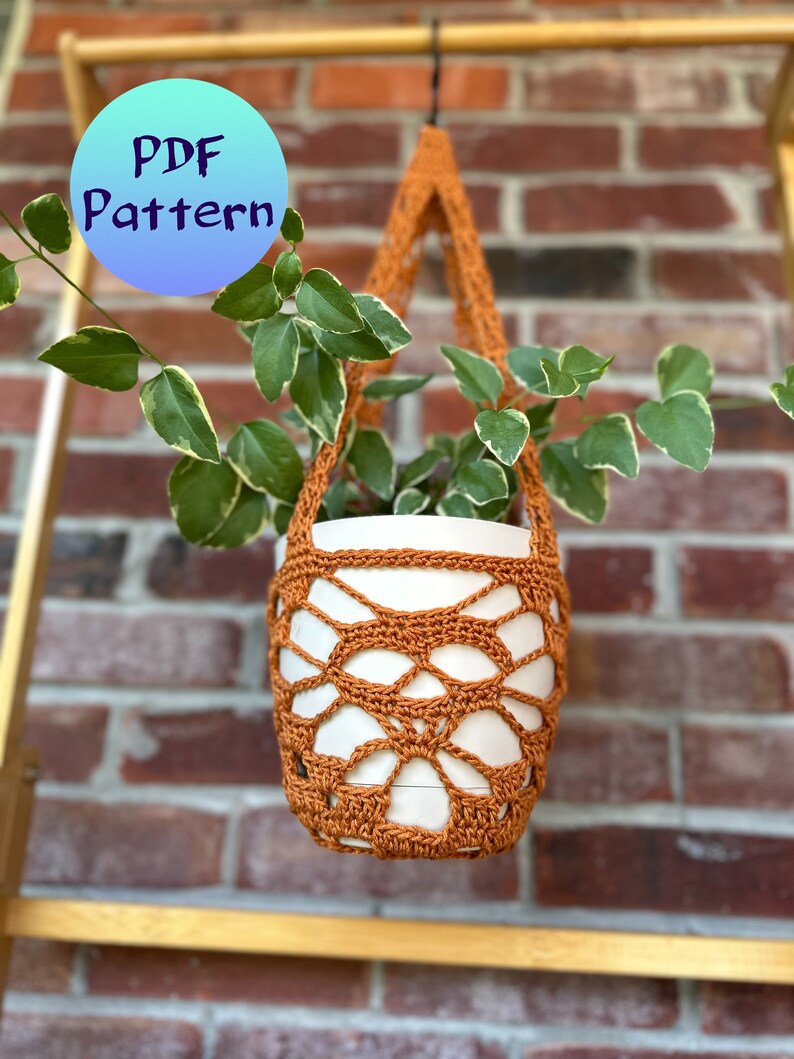 May include: A crocheted plant hanger in a burnt orange color hangs from a hook against a brick wall. The hanger is holding a white pot with a green plant.