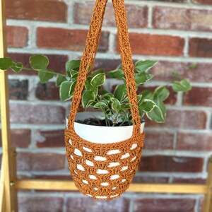 May include: A brown crocheted hanging planter with a white pot and a green plant inside. The planter is hanging from a wooden shelf.