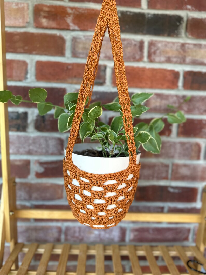 May include: A brown crocheted hanging planter with a white pot and a green plant inside. The planter is hanging from a wooden shelf.