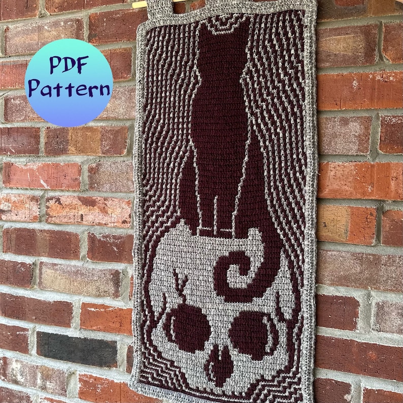 May include: A crocheted wall hanging featuring a black cat standing on top of a skull. The skull is gray with burgundy accents. The background is a gray and white striped pattern.