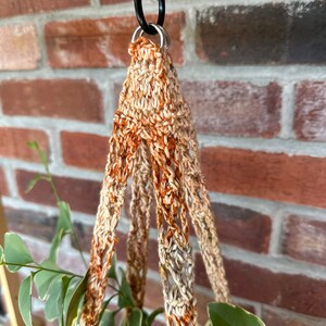 May include: A close-up of a macrame plant hanger with a brown and beige color scheme. The hanger is made of crocheted cord and has a loop at the top for hanging.