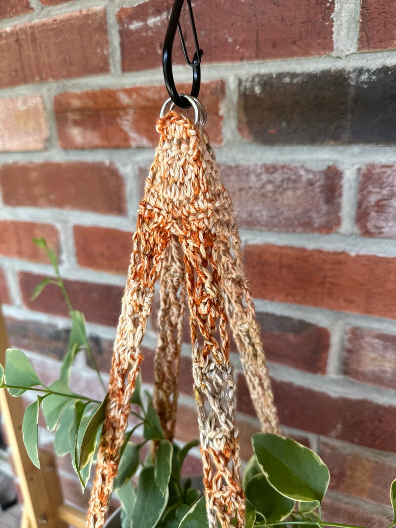 May include: A close-up of a macrame plant hanger with a brown and beige color scheme. The hanger is made of crocheted cord and has a loop at the top for hanging.