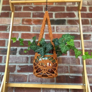 May include: A brown crocheted plant hanger with a green potted plant hanging from a wooden rack against a brick wall.