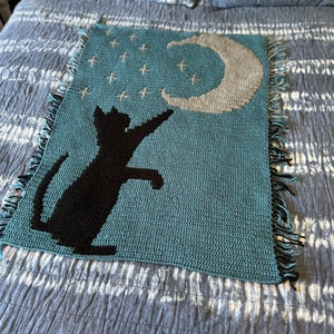 May include: A teal blue crocheted blanket featuring a black cat silhouette reaching up to a crescent moon with white stars on a dark blue background.