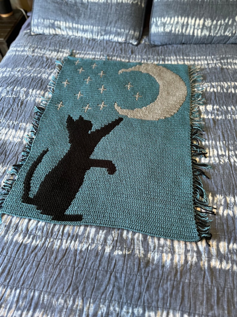 May include: A teal blue crocheted blanket featuring a black cat silhouette reaching up to a crescent moon with white stars on a dark blue background.