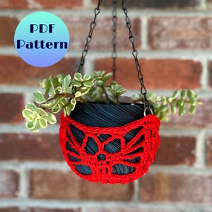 Pode incluir: Um vaso de plantas suspenso preto com um design de crochê vermelho, contendo uma planta verde com folhas verde-amareladas. O vaso é suspenso por uma corrente preta contra uma parede de tijolos. O texto "PDF Pattern" está em um círculo azul.