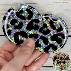 May include: Two round black and silver glitter coasters with a leopard print design. The coasters are made of resin and have a glossy finish.