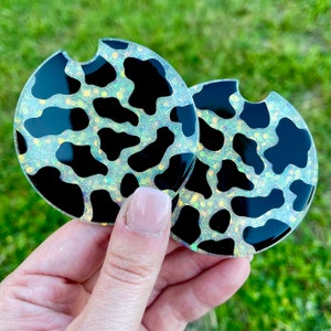 May include: Two round black and white cow print coasters with a glittery finish. The coasters are made of a clear material and have a hole in the center for a cup.