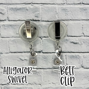 May include: Two clear plastic retractable badge holders with metal clips. The holder on the left has an alligator swivel clip and the holder on the right has a belt clip. Both holders are hanging from a white brick wall.