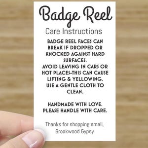 May include: White card with black text that reads "Badge Reel Care Instructions". The text explains how to care for a badge reel, including avoiding heat and using a gentle cloth to clean. The card also says "Handmade with love. Please handle with care." and "Thanks for shopping small, Brookwood Gypsy."