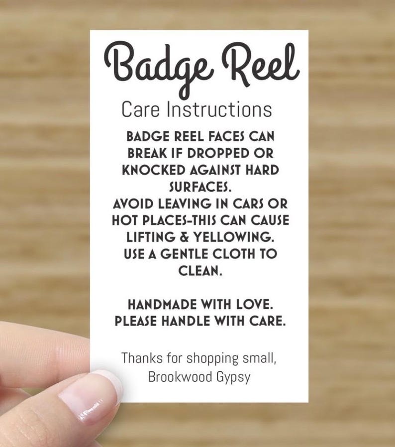 May include: White card with black text that reads "Badge Reel Care Instructions". The text explains how to care for a badge reel, including avoiding heat and using a gentle cloth to clean. The card also says "Handmade with love. Please handle with care." and "Thanks for shopping small, Brookwood Gypsy."