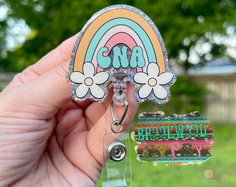 May include: A badge reel with a rainbow design in pastel colors, featuring the letters "CNA" in turquoise. Two white flowers with yellow centers are at the bottom. The reel has a clear plastic strap and a metal clip.