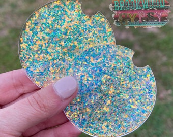 May include: Two iridescent glitter-filled car coasters with a clear plastic backing. The coasters are round and have a cutout for a cup.