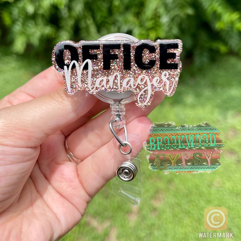 May include: A pink and black glitter badge holder with the text "Office Manager" on it. The badge holder is attached to a retractable badge reel.