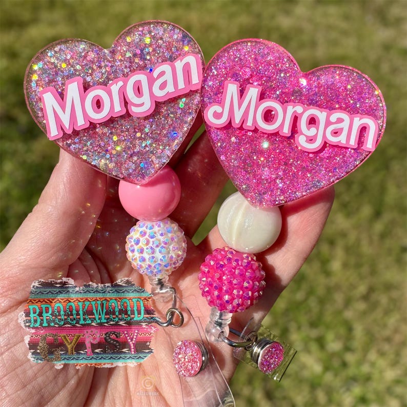 May include: Pink glitter heart shaped badge holder with the name "Morgan" on it. The badge holder has a pink, white, and iridescent bead charm. The badge holder is attached to a clear retractable badge reel.