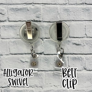 May include: Two clear plastic retractable badge holders with metal clips. The holder on the left has an alligator swivel clip and the holder on the right has a belt clip. Both holders are hanging from a white brick wall.