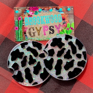 May include: Two white and black cow print car coasters with iridescent glitter. The coasters are round and have a cut-out for a cup. The coasters are on a red and black plaid background. The text "Brookwood Gypsy" is visible in the background.