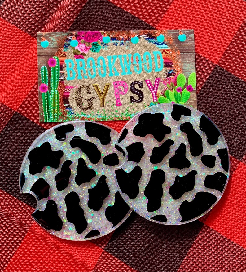 May include: Two white and black cow print car coasters with iridescent glitter. The coasters are round and have a cut-out for a cup. The coasters are on a red and black plaid background. The text "Brookwood Gypsy" is visible in the background.