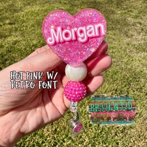 May include: Hot pink glitter heart shaped badge holder with the name "Morgan" in a retro font. The holder has a white pearl and a pink rhinestone bead.