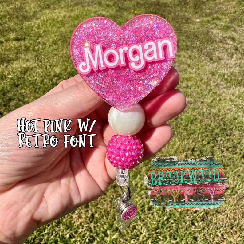 May include: Hot pink glitter heart shaped badge holder with the name "Morgan" in a retro font. The holder has a white pearl and a pink rhinestone bead.