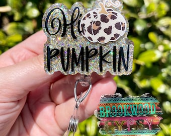 以下が含まれることがあります： 黒い筆記体で「Hi PUMPKIN」と書かれたキラキラ光るバッジリール。ヒョウ柄のデザインのカボチャがトッピングされています。リールには、カラフルな南西風のパターンで「BROOKWOOD Y'S」と書かれた小さなタグも付いています。