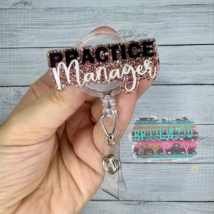 May include: A pink glitter badge holder with a black and white text overlay that reads "PRACTICE Manager". The badge holder is attached to a clear retractable badge reel.